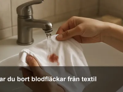 Hand som spolar bort en blodfläck från en vit T‑shirt under kranen med kallt vatten i en handfat.